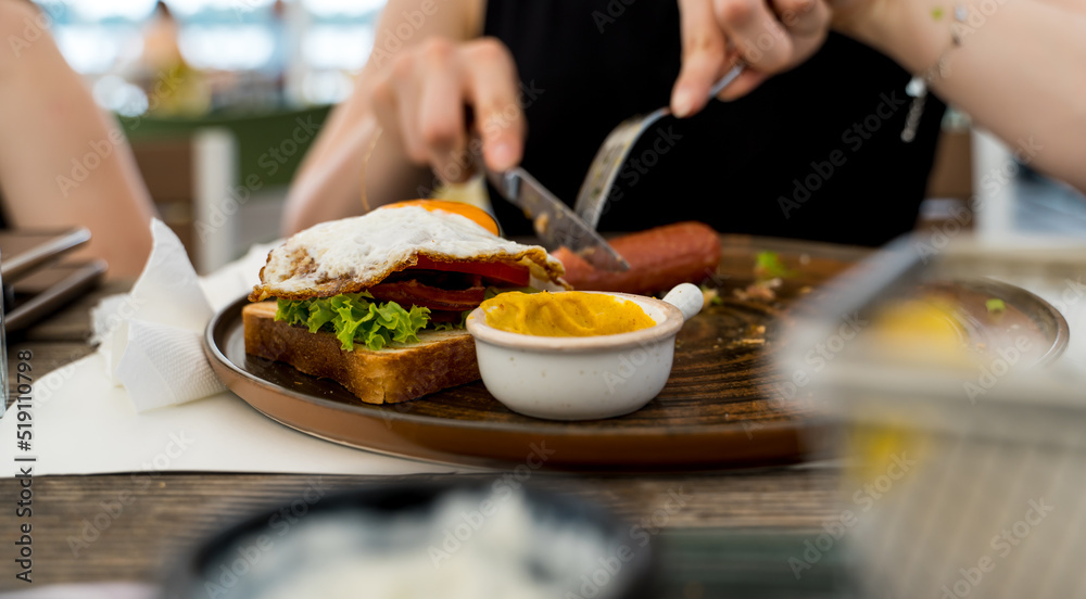 A close up girl is eating healthy English breakfast during the day in a restaurant cutting sausage and sandwich 