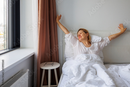 An elderly woman stretches sweetly in her bed after sleeping.