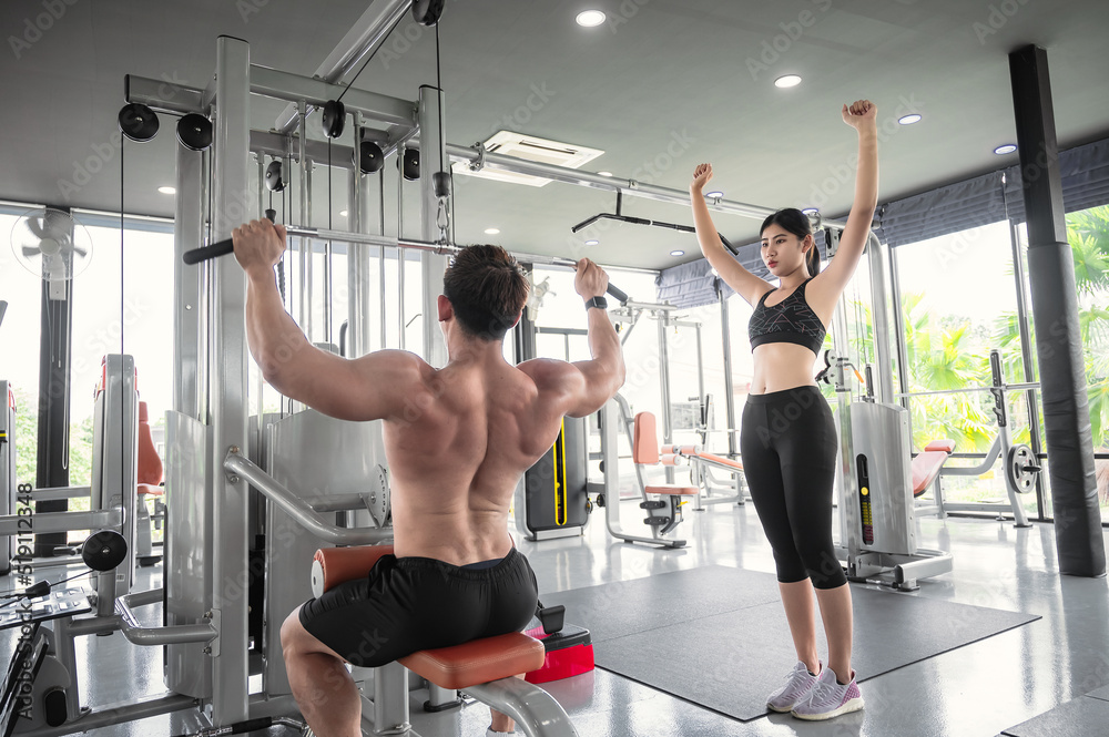 Fototapeta premium Young Asian bodybuilder trainer showing correct use of exercise equipment for Asian women to follow, in fitness room. Asian man trainer is teaching Asian women correct use of exercise equipment in gym