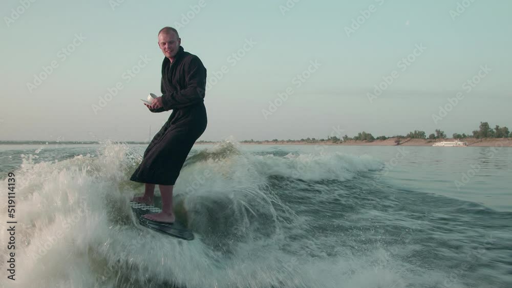 A surfer jumping on a wakeboard in a terry dressing gown with a cup of tea or coffee in his hands. An experienced wakeboarder sprays water drops into the camera. A surfer jumping on a wakeboard in a terry dressing gown with a cup of tea or coffee in his hands. An experienced wakeboarder sprays water drops into the camera.