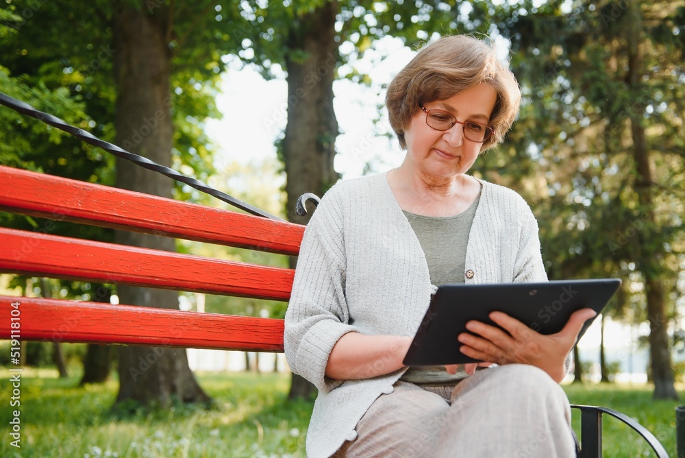 Fototapeta premium Elegant elderly woman in the shirt is sitting on the bench in a park on a warm day