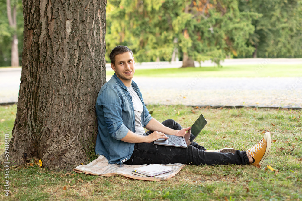 Obraz premium Young man with laptop rest in grass at university campus. Business man sit on lawn work outdoors on computer in a park. Student man on lesson with laptop. education and remote working concept