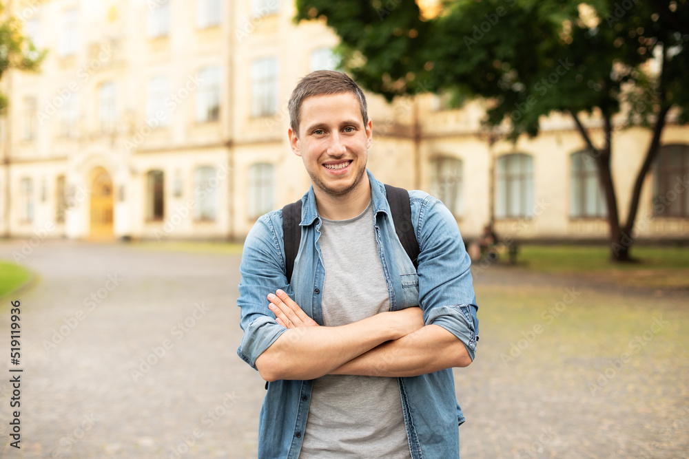 Successful student is standing with backpack near campus on the university background. Smiling young man with bag standing in park. Education, university, college, studying, course concept
