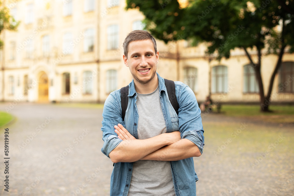 Fototapeta premium Successful student is standing with backpack near campus on the university background. Smiling young man with bag standing in park. Education, university, college, studying, course concept 