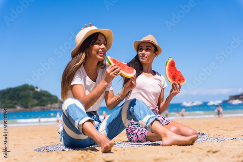 Two sisters in summer on the beach eating a watermelon, on vacation with the sea in the background