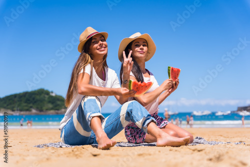 Two sisters in summer on the beach eating a watermelon, on vacation with the sea in the background
