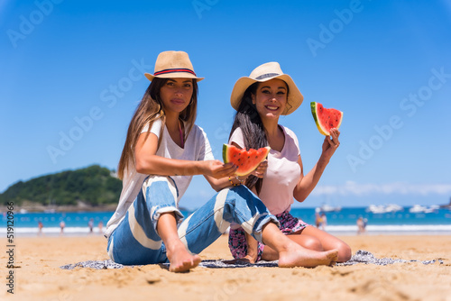 Two friends in summer on the beach eating a watermelon, on vacation with the sea in the background