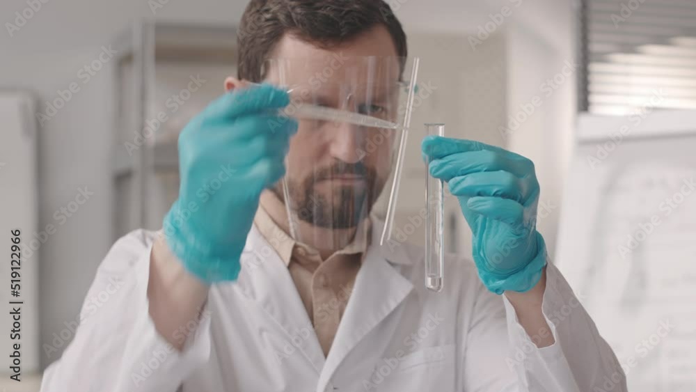Chest up slowmo of serious Caucasian chemist in face shield and lab coat conducting experiment in lab adding hydrogen peroxide and some black powder in test tube and watching chemical reaction