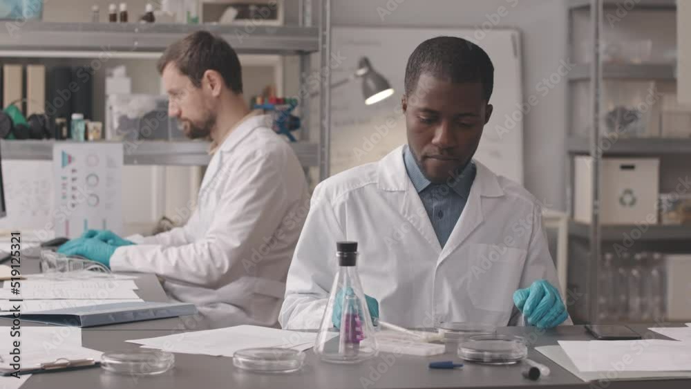 Medium slowmo of young African American chemist dropping some liquid substance in petri dish and taking picture of chemical reaction on smartphone, working in lab with assistant Medium slowmo of young African American chemist dropping some liquid substance in petri dish and taking picture of chemical reaction on smartphone, working in lab with assistant