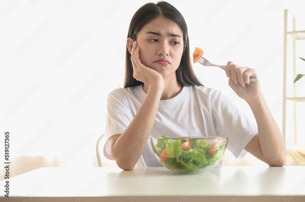 Beautiful young Asian woman feeling boring while eating fresh vegetable. Woman do not want to eat vegetables because of taste of vegetable or dislike dieting. Beautiful young Asian woman feeling boring while eating fresh vegetable. Woman do not want to eat vegetables because of taste of vegetable or dislike dieting.