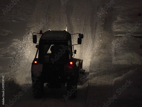 Wheeled tractor cleaning a snow with snowplow on snowy road at winter night after snowfall with headlight