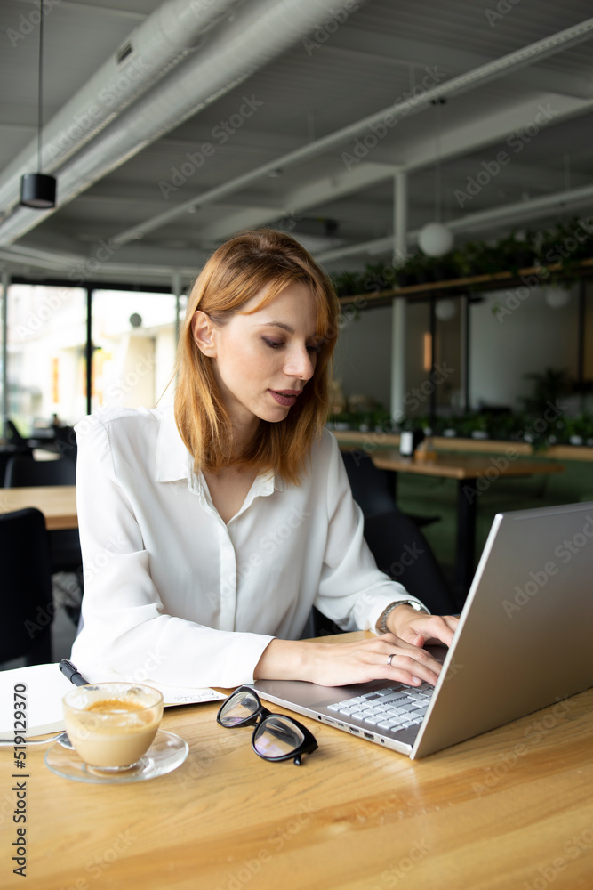 White slim woman with red hair in white shirt sitting at the table and tipping in the grey laptop with a cup of coffee on the table