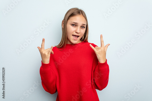 Young caucasian girl isolated on blue background showing a horns gesture as a revolution concept.