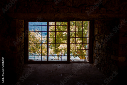 View through the bars of an old prison cell. Outside you can see the sandy beach, on which is a large willow tree.