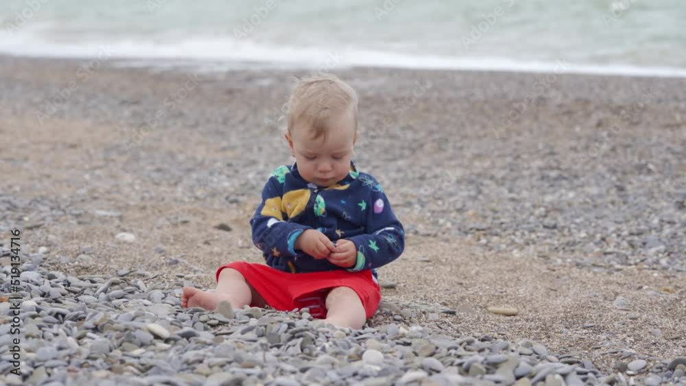 Kid sits on a windy beach near the sea, 1.5 year old toddler child is playing with pebbles in summer. Kid sits on a windy beach near the sea, 1.5 year old toddler child is playing with pebbles in summer.