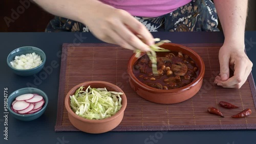 Decorating vegan Pozole Rojo with shredded ice lettuce, radish and white onion. Mexican dish with hominy, shredded jackfruit, button mushrooms and dried chilies: guaillo, ancho, pasilla. photo