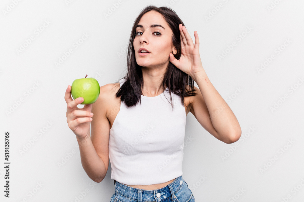 Young caucasian woman holding an apple isolated on white background trying to listening a gossip. Young caucasian woman holding an apple isolated on white background trying to listening a gossip.