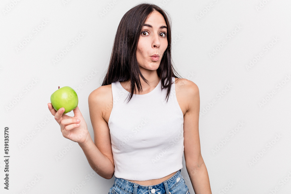 Young caucasian woman holding an apple isolated on white background shrugs shoulders and open eyes confused. Young caucasian woman holding an apple isolated on white background shrugs shoulders and open eyes confused.