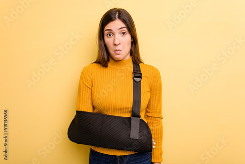 Young caucasian woman with broke arm isolated on yellow background shrugs shoulders and open eyes confused. photo