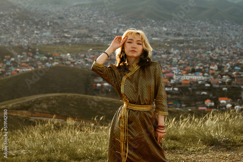 Young Mongolian woman wears traditional golden deel dress. Ulaanbaatar in background at sunset.