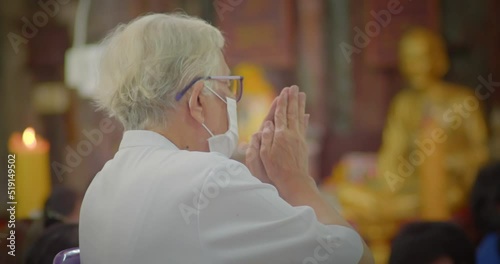 Slow motion scene of an Asian lady who is an elderly has white hair, wearing glasses and a mask to prevent COVID-19 , sitting and hands clasping at the merit making ceremony in the temple. photo