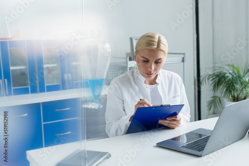 Scientist writing on clipboard near laptop and flask in lab. © LIGHTFIELD STUDIOS