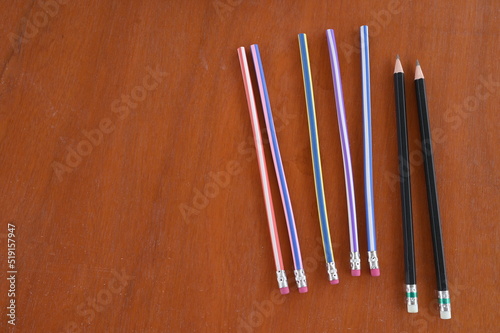 5 unsharpened pencils of different colors but with the same pink eraser and 2 black sharpened pencils with white erasers all placed on a wooden table. Two pencils were sharpened with a sharpener. 