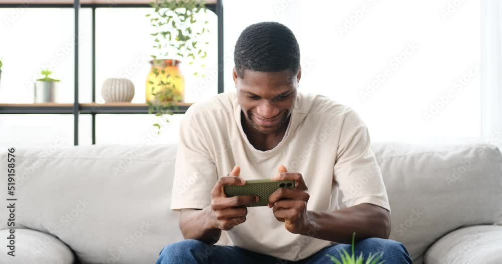 African american man playing game using mobile phone while sitting on sofa at home African american man playing game using mobile phone while sitting on sofa at home