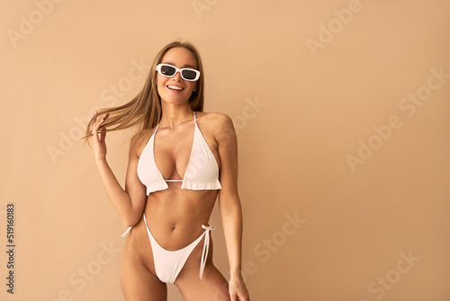 A tanned young woman in a white swimsuit and white sunglasses laughs on a beige background.
