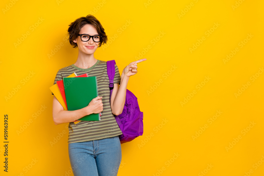 Photo of pretty sweet girl dressed green t-shirt eyewear rucksack holding book pointing empty space isolated yellow color background Photo of pretty sweet girl dressed green t-shirt eyewear rucksack holding book pointing empty space isolated yellow color background