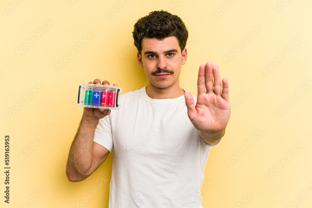 Young caucasian man holding batteries isolated on yellow background standing with outstretched hand showing stop sign, preventing you. Young caucasian man holding batteries isolated on yellow background standing with outstretched hand showing stop sign, preventing you.
