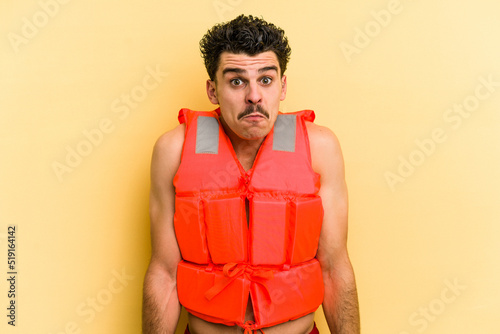 Young caucasian man wearing life jacket isolated on yellow background shrugs shoulders and open eyes confused. © Asier