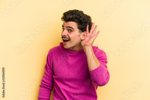 Young caucasian man isolated on yellow background trying to listening a gossip.