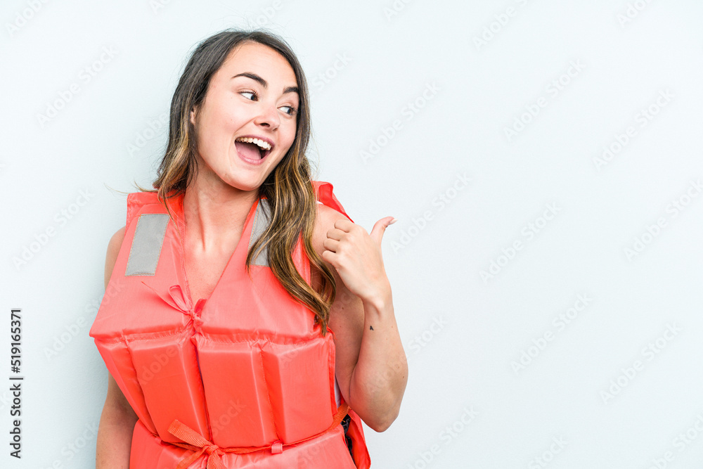 Young caucasian woman wearing life jacket isolated on blue background points with thumb finger away, laughing and carefree. Young caucasian woman wearing life jacket isolated on blue background points with thumb finger away, laughing and carefree.