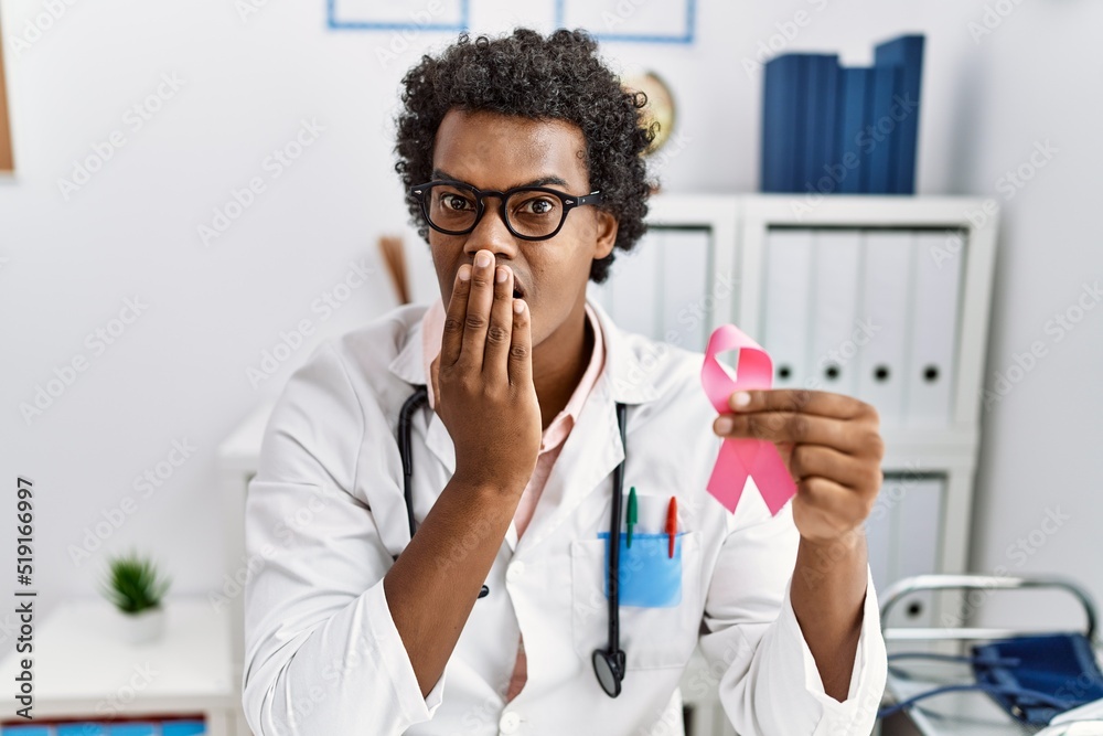 African doctor man holding pink cancer ribbon covering mouth with hand, shocked and afraid for mistake. surprised expression African doctor man holding pink cancer ribbon covering mouth with hand, shocked and afraid for mistake. surprised expression