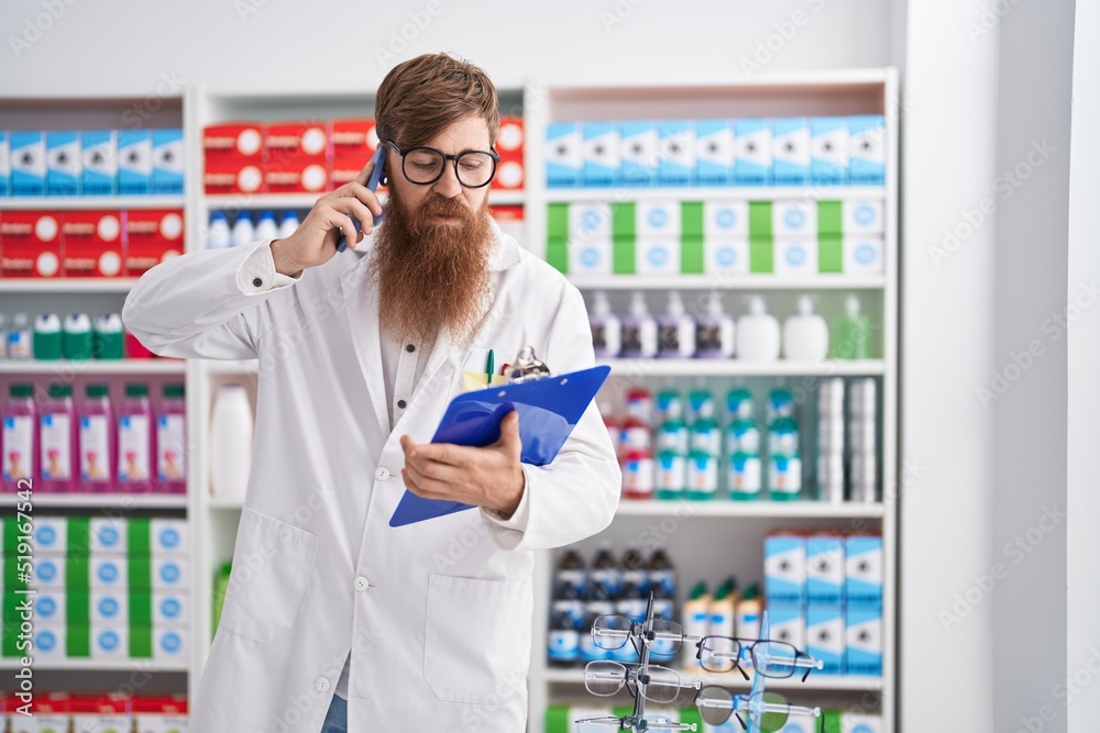 Young redhead man pharmacist talking on smartphone reading document at pharmacy