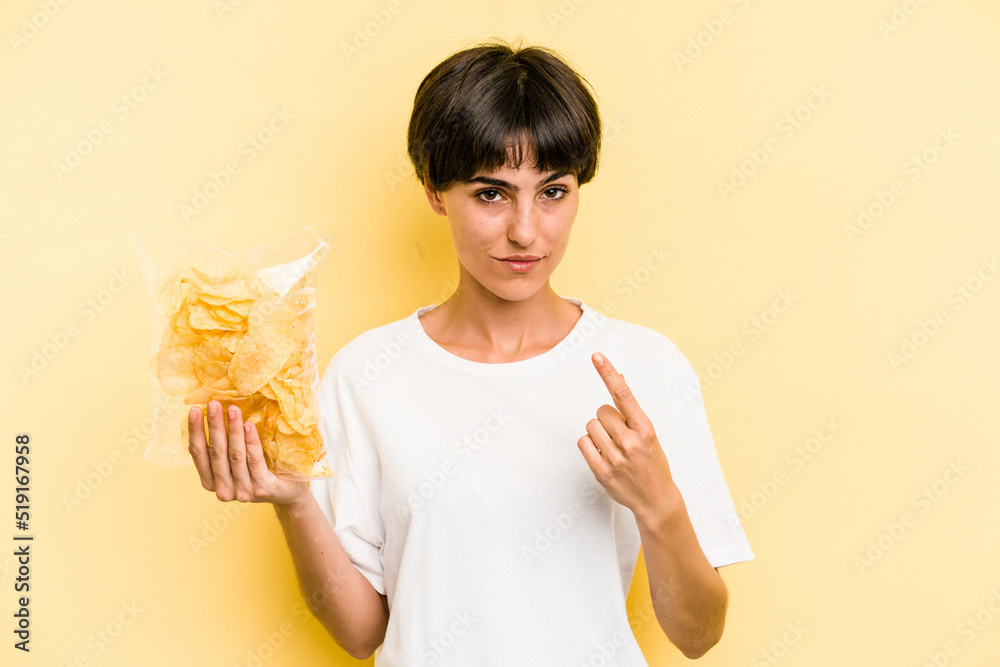 Young caucasian man holding crisps isolated on yellow background pointing with finger at you as if inviting come closer. Young caucasian man holding crisps isolated on yellow background pointing with finger at you as if inviting come closer.