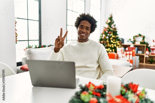 Young african american man using laptop sitting on the table by christmas tree showing and pointing up with fingers number two while smiling confident and happy.