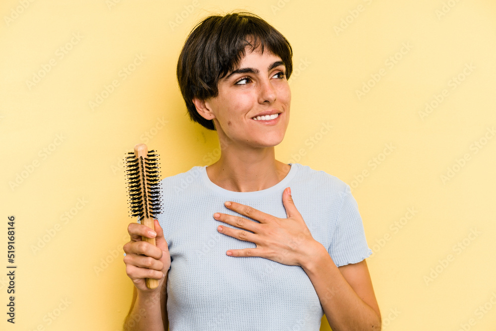 Young caucasian woman holding a brush hair isolated on yellow background laughs out loudly keeping hand on chest. Young caucasian woman holding a brush hair isolated on yellow background laughs out loudly keeping hand on chest.