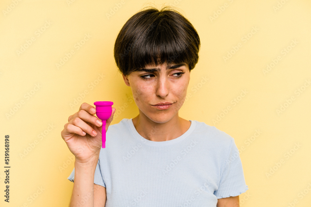 Young caucasian woman holding a menstrual cup isolated on yellow background confused, feels doubtful and unsure. Young caucasian woman holding a menstrual cup isolated on yellow background confused, feels doubtful and unsure.
