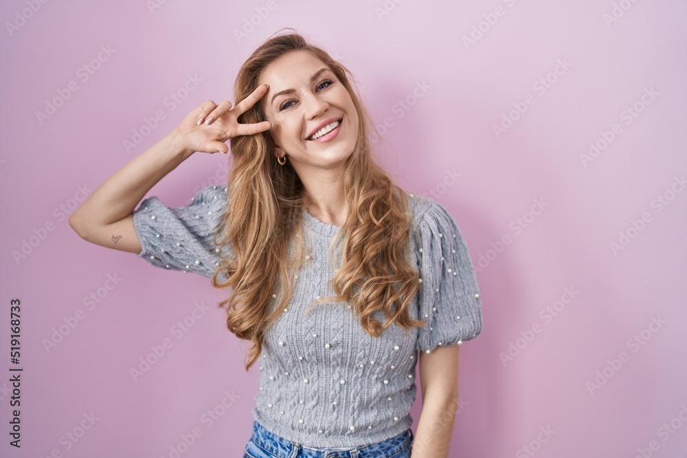 Beautiful blonde woman standing over pink background doing peace symbol with fingers over face, smiling cheerful showing victory Beautiful blonde woman standing over pink background doing peace symbol with fingers over face, smiling cheerful showing victory