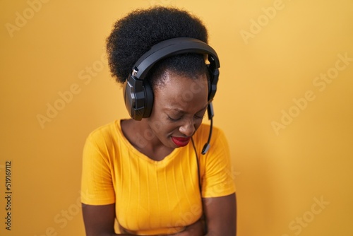 African woman with curly hair standing over yellow background wearing headphones with hand on stomach because nausea, painful disease feeling unwell. ache concept.