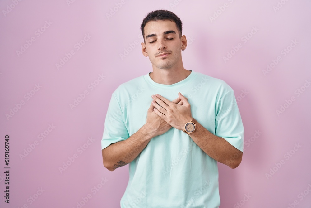 Handsome hispanic man standing over pink background smiling with hands on chest with closed eyes and grateful gesture on face. health concept. Handsome hispanic man standing over pink background smiling with hands on chest with closed eyes and grateful gesture on face. health concept.
