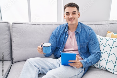 Young hispanic man drinking coffee using touchpad at home