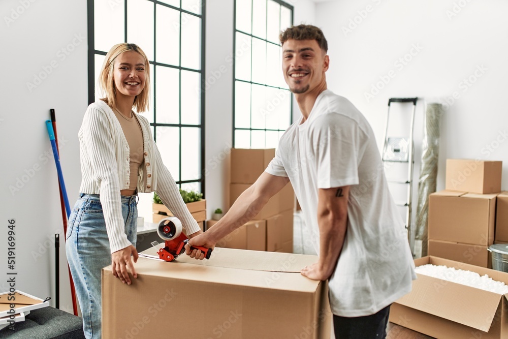 Obraz premium Young caucasian couple smiling happy closing box using packing tape at new home.
