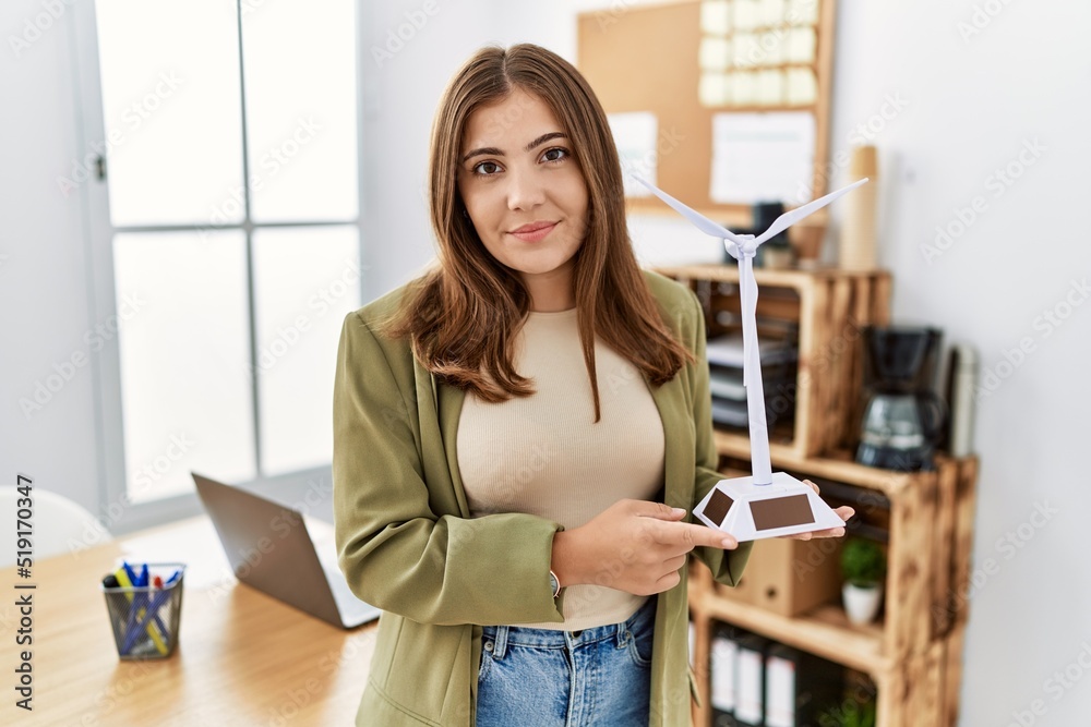 Young brunette woman holding solar windmill for renewable electricity at the office thinking attitude and sober expression looking self confident Young brunette woman holding solar windmill for renewable electricity at the office thinking attitude and sober expression looking self confident