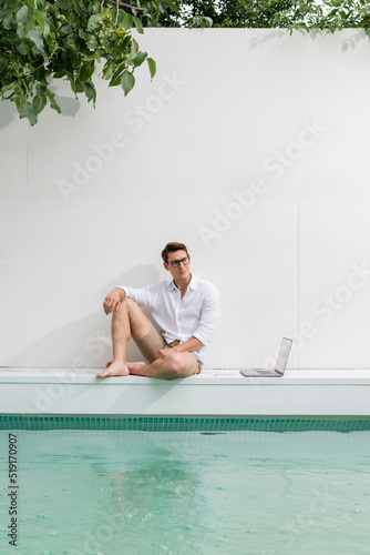 full length of barefoot man in eyeglasses sitting at poolside near white wall and laptop.