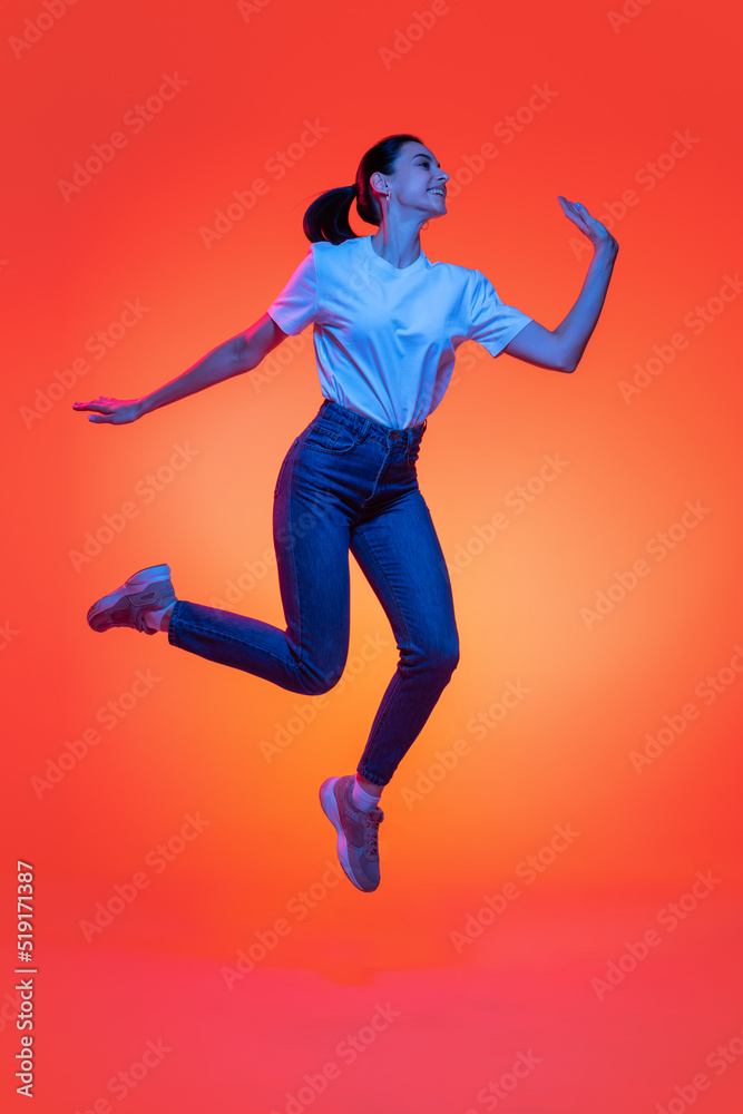 Full-length portrait of young pretty girl, student in casual style clothes isolated on orange color background in neon light. Concept of beauty, fashion, emotions