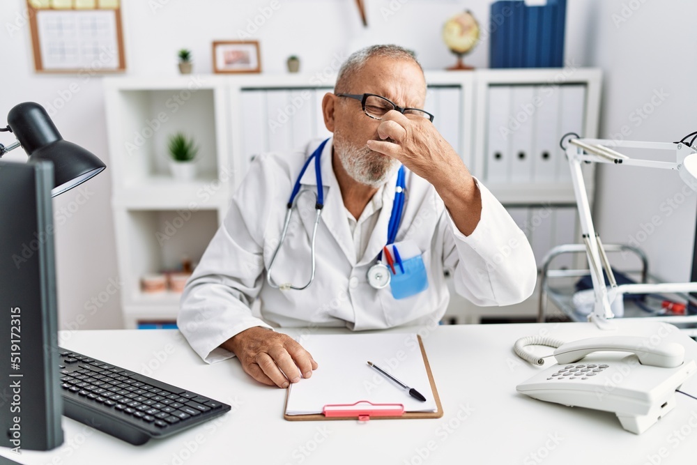 Mature doctor man at the clinic smelling something stinky and disgusting, intolerable smell, holding breath with fingers on nose. bad smell