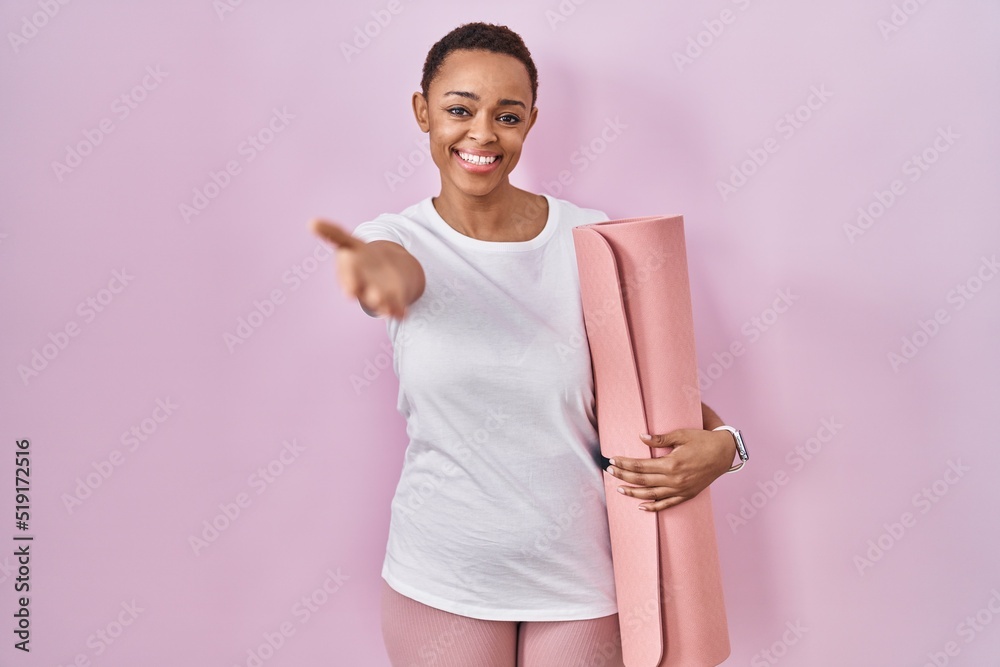 Beautiful african american woman holding yoga mat smiling cheerful offering palm hand giving assistance and acceptance.